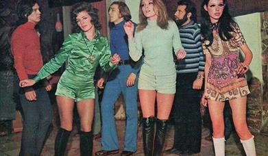 A photograph from the 1960s featuring three young women in jumpsuits and three young men in polo neck shirts and flares dancing indoors