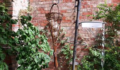 A woven wicker bird feeder hanging in a garden. The bird feeder contains several fat balls.