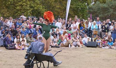 Two performers are outdoors in front of a large audience. One performer is dancing, wearing a bright green leotard and an oversized, colourful flower-