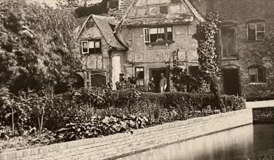 A black and white photograph of a Victorian man and woman standing outside a timber framed cottage beside a river