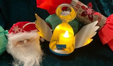 A brightly coloured paper Christmas cracker, a metal tea light in the shape of an angel, and a fabric decoration depicting Santa's face