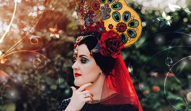 A woman in a traditional Ingusheti headdress in bright red and yellow embroidered with green peacock feathers standing outside in front of green folia