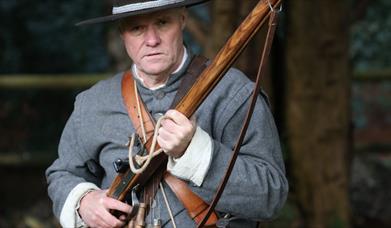 A photograph of an English Civil War re-enactor wearing a wide-brimmed felt hat and a bandoleer and holding a rifle