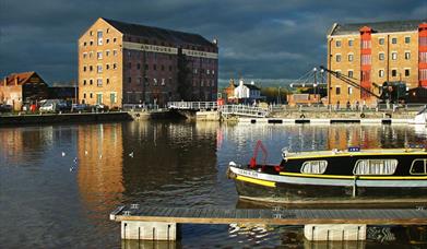 The large brown warehouse buildings of Gloucester Docks bordering the water of the canal, with narrowboats in the foreground.