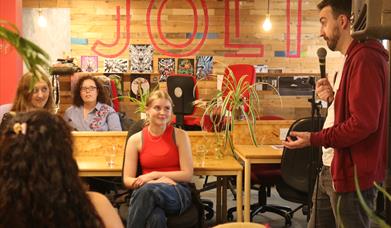A diverse audience of all ages watches a dynamic PechaKucha-style talk and speaker presentation at Jolt Studios and event space, part of a creative co