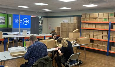 People working with archive boxes in a records room with shelves and tables.