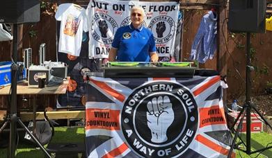 A man in a blue polo shirt standing behind a large record deck with a banner hung over it that reads; "Northern Soul: way of life"