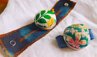 Two pin cushions made from multicoloured floral patterned fabric on blue demin wrist straps.