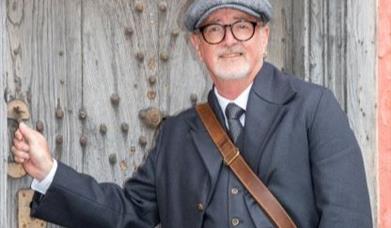 A smiling man in an Edwardian costume with flat cap and waistcoat opening a large old oak door