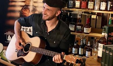 A young man wearing a flat cap sitting on a stool in a whiskey bar playing guitar