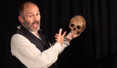 A man in a white shirt and black waistcoat holding a skull and pointing at it in mid speech