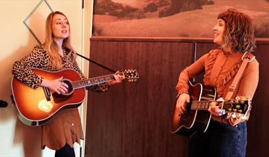 Two young women standing indoors playing acoustic guitars and singing while looking at each other