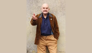 A man standing against a beige pebble dash wall with his hand out in mid speech