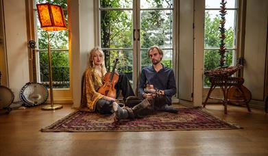 A young woman holding a viola sitting next to a young man holding a violin on an Afghan rug on the floor.