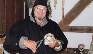 A man in a black Tudor smock and hat grinning humorously while holding a model of a skull and a pair of tooth pulling pliers