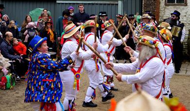 A group of Morris dancers in white outfits with bells and sticks dancing in front of a crowd of people in winter outdoors