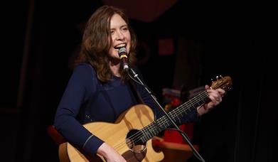 A woman with shoulder length dark brown hair singing and playing acoustic guitar