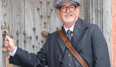 A male tour guide dressed as an Edwardian undertaker opening the wooden front door of a Tudor house.
