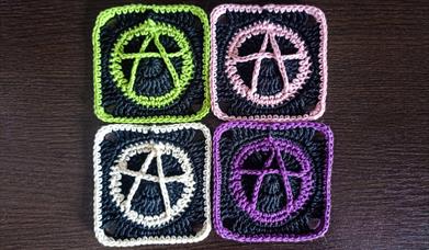 Four crocheted "granny squares" with the anarchy symbol sewn into them