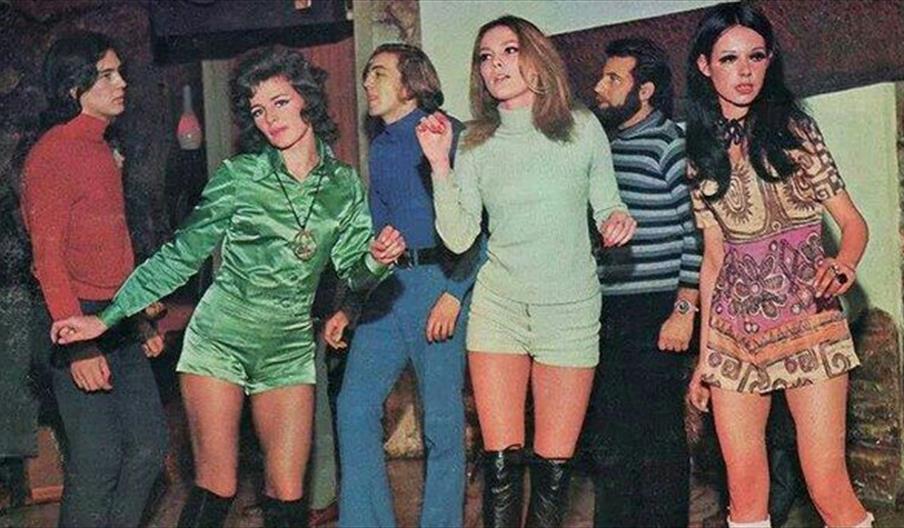 A photograph from the 1960s featuring three young women in jumpsuits and three young men in polo neck shirts and flares dancing indoors