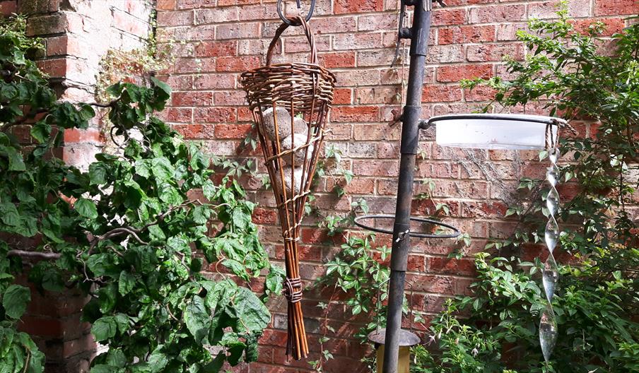 A woven wicker bird feeder hanging in a garden. The bird feeder contains several fat balls.