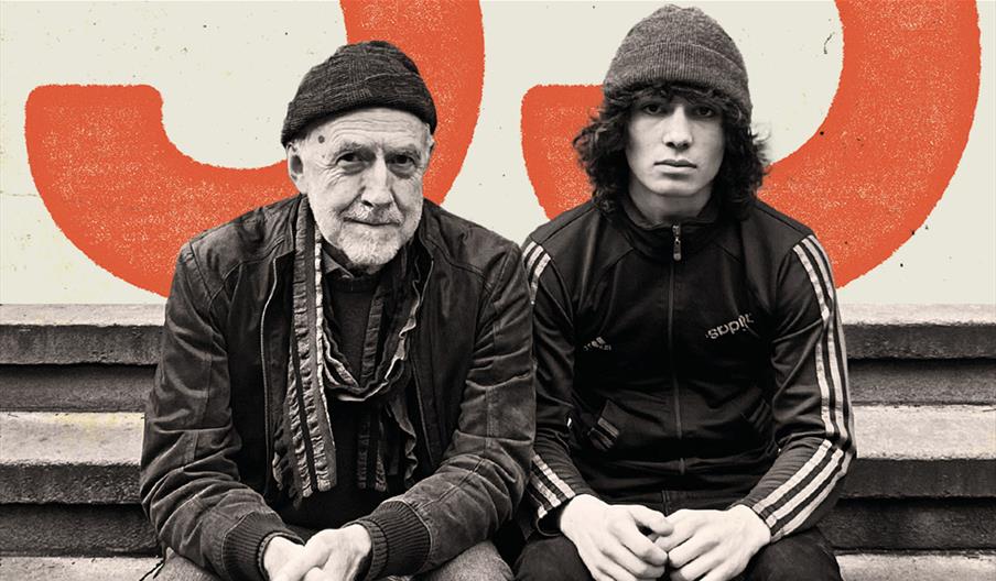 Two performers, one young and one older sat together in front of a large orange 55.