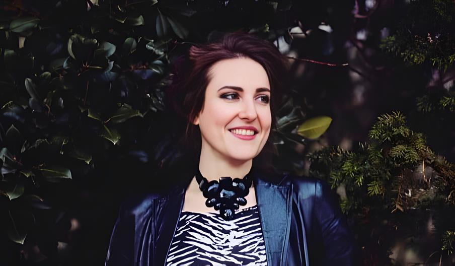 A smiling young woman with dark reddish brown hair standing in front of evergreen foliage outdoors.