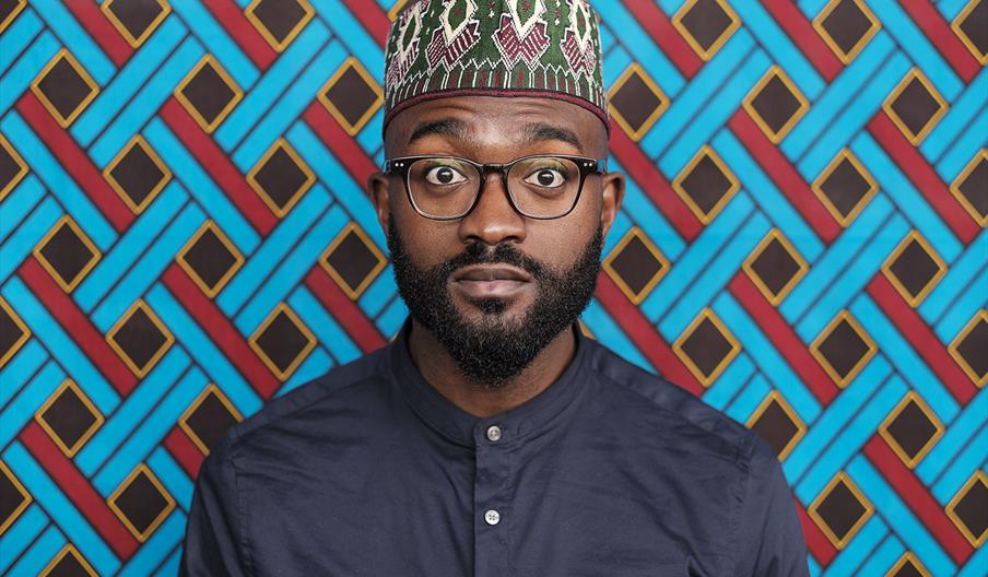 Inua stands against a brightly coloured, patterned backdrop, wearing glasses, a blue shirt and a traditional patterned hat.