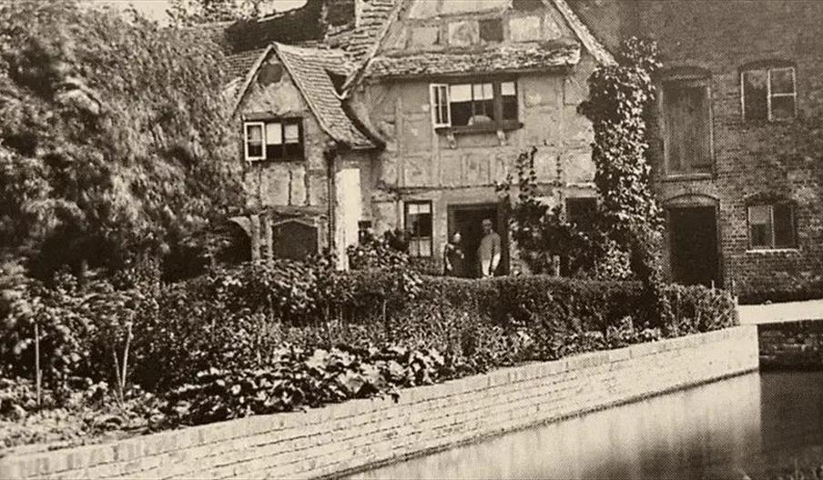 A black and white photograph of a Victorian man and woman standing outside a timber framed cottage beside a river