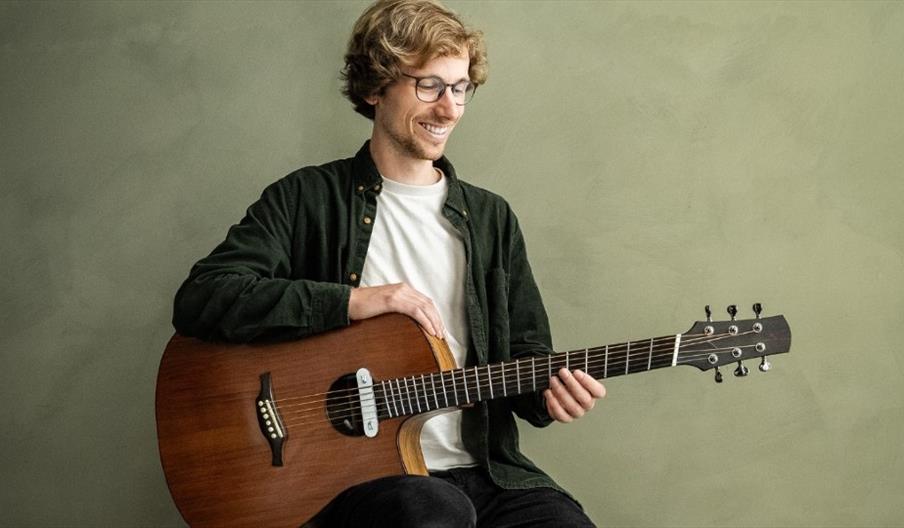 A smiling young man with wavy light brown hair sitting on a stool holding an acoustic guitar