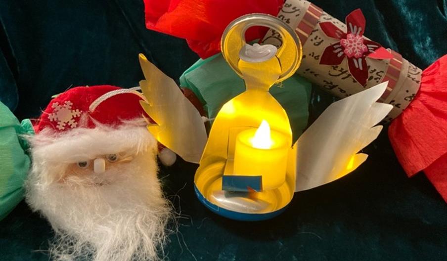 A brightly coloured paper Christmas cracker, a metal tea light in the shape of an angel, and a fabric decoration depicting Santa's face