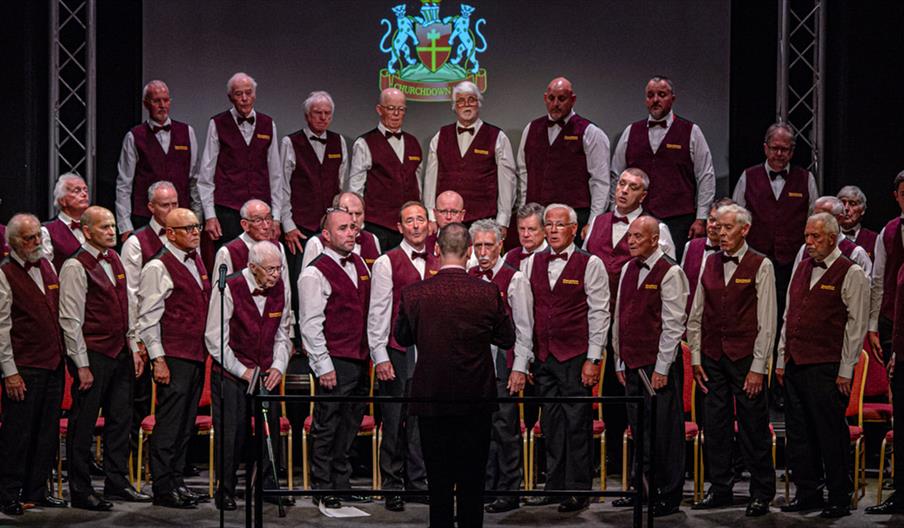 A male choir performing on stage, dressed in white shirts, maroon vests, and black bow ties, with a conductor in front and a crest featuring two lions
