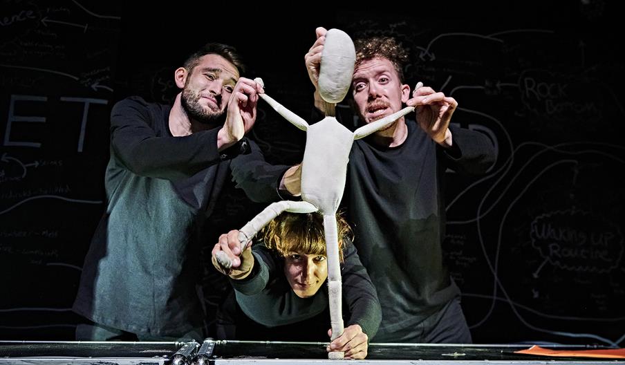 Fred, a two-foot-tall white cloth puppet, is stood dramatically with his arms and one leg stuck out, lit up against a darker background. His three pup