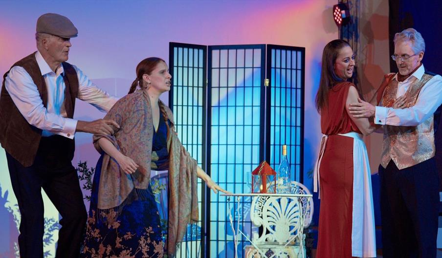 Two male singers in smart shirts and waistcoats and two female singers in long dresses enacting a dramatic scene on stage indoors.