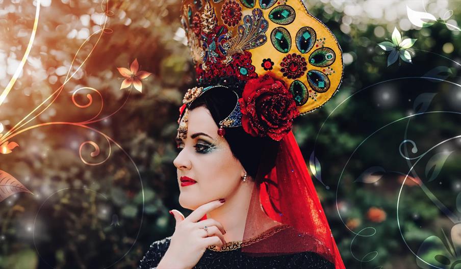 A woman in a traditional Ingusheti headdress in bright red and yellow embroidered with green peacock feathers standing outside in front of green folia