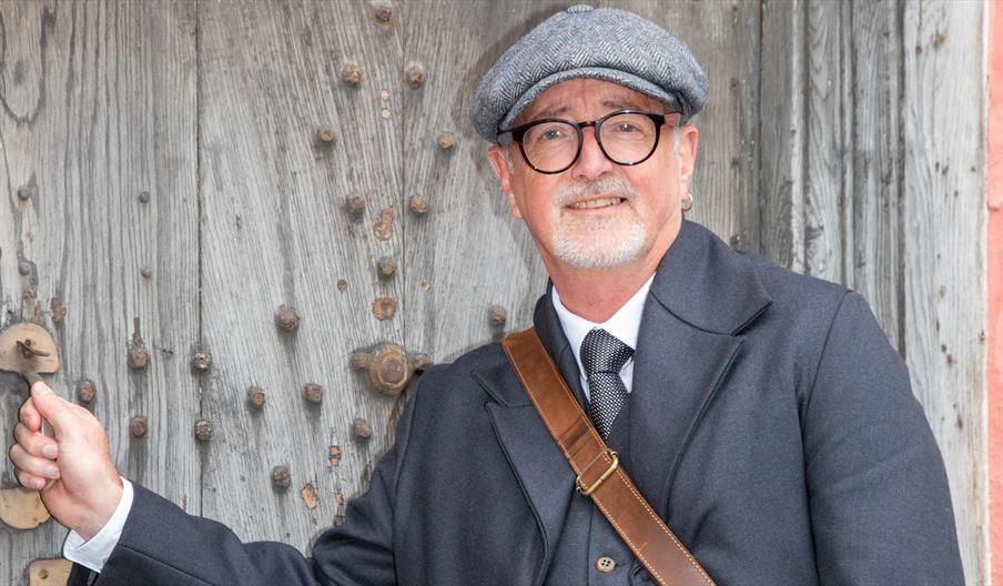A smiling man in an Edwardian costume with flat cap and waistcoat opening a large old oak door