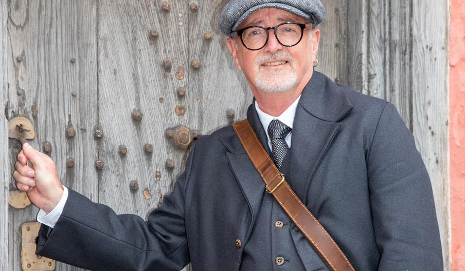 A male tour guide dressed as an Edwardian undertaker opening the wooden front door of a Tudor house.