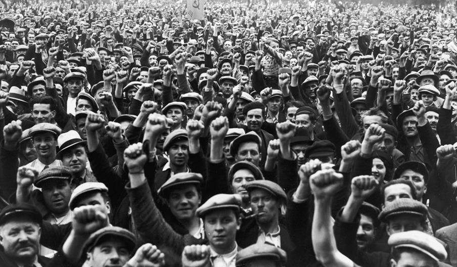A black and white photograph of a crowd of people outdoors raising their fists in the air. They are wearing shirts, jackets, flat caps and trilby hats