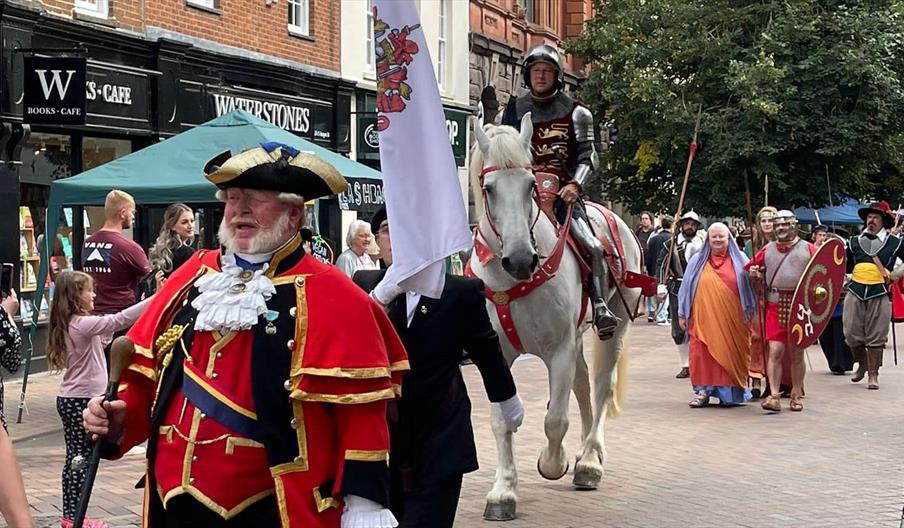 A procession of people in historical costumes walking up Westgate Street in Gloucester, including a medieval knight on horseback and an 18th century m