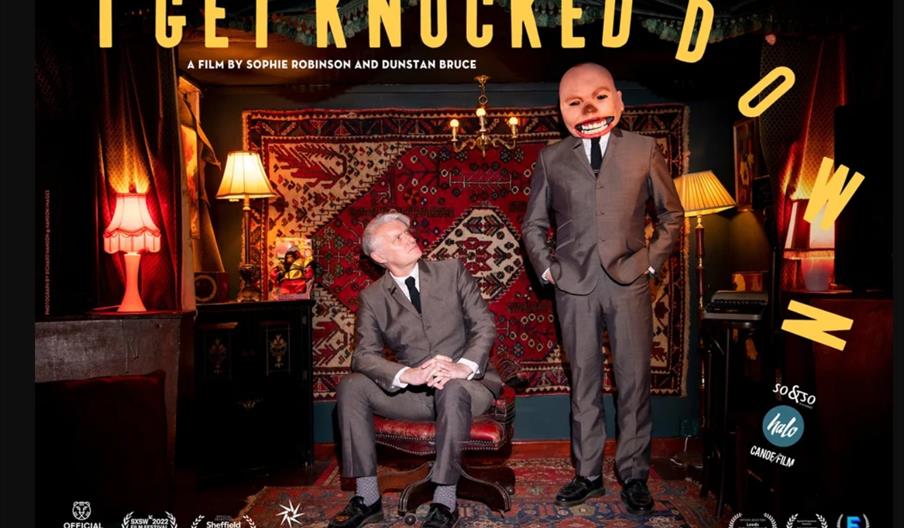 The film poster for "I Get Knocked Down", featuring Dunstan Bruce, frontman of Anarchist band humbawamba, sitting on a chair in a living room and look