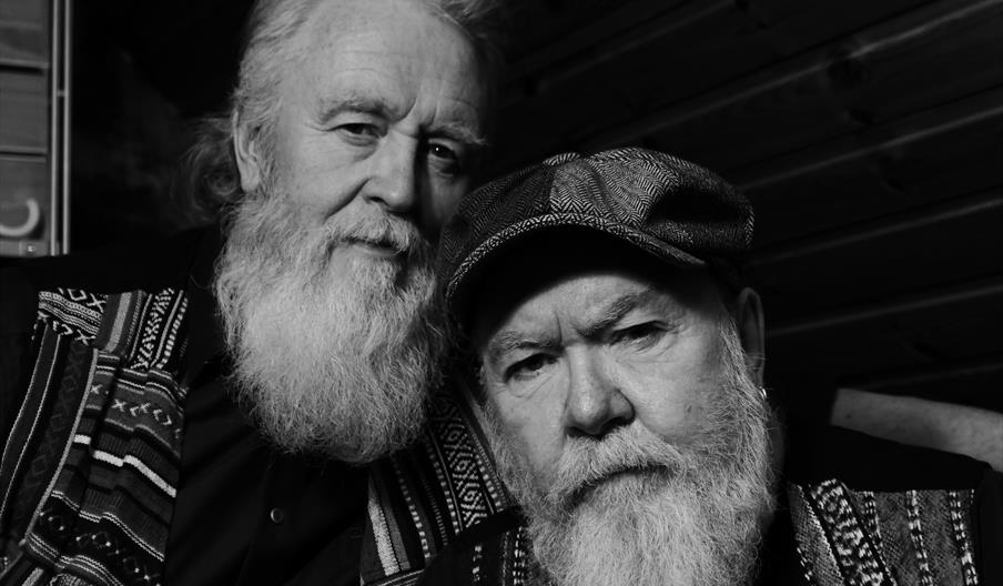A black and white photograph of two men with white beards sitting indoors in front of a wood panelled wall.