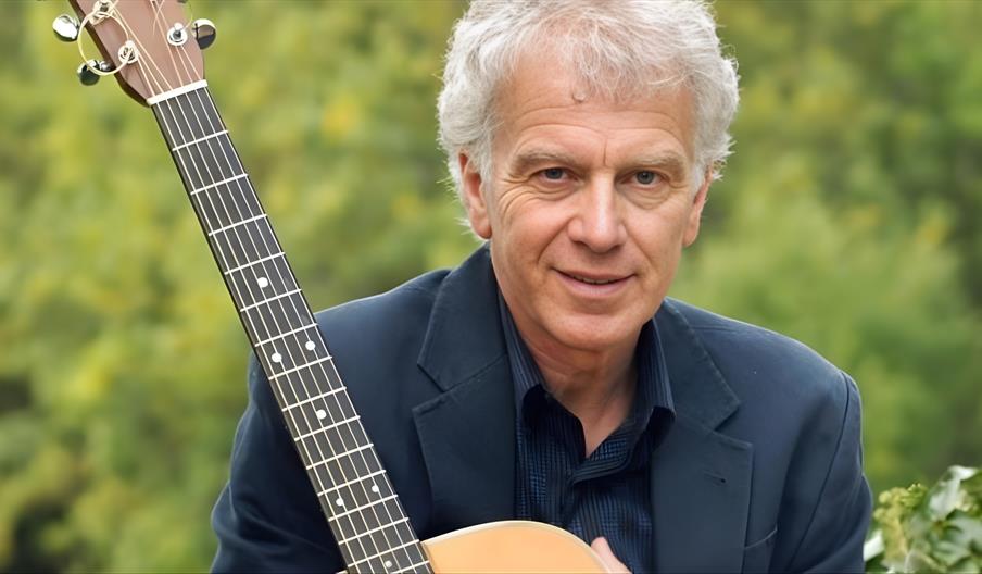 A smiling man with curly white hair holding an acoustic guitar.