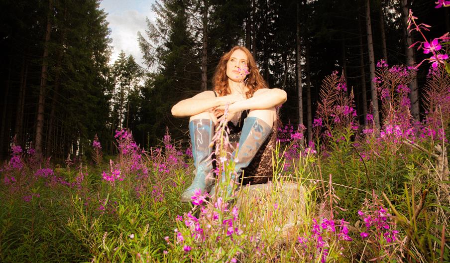 A photo of a woman sitting in a field of purple flowers surrounding her and tall trees behind her. She is wearing blue knee-high boots and a black lac