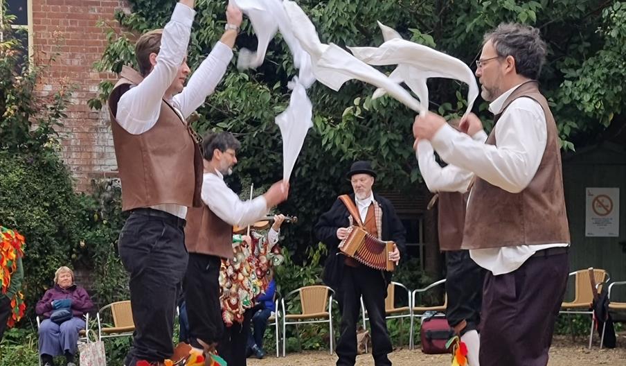 Four Morris dancers in brown leather waistcoats leaping into the air waving white handkerchiefs, with a man playing accordion and a woman playing viol