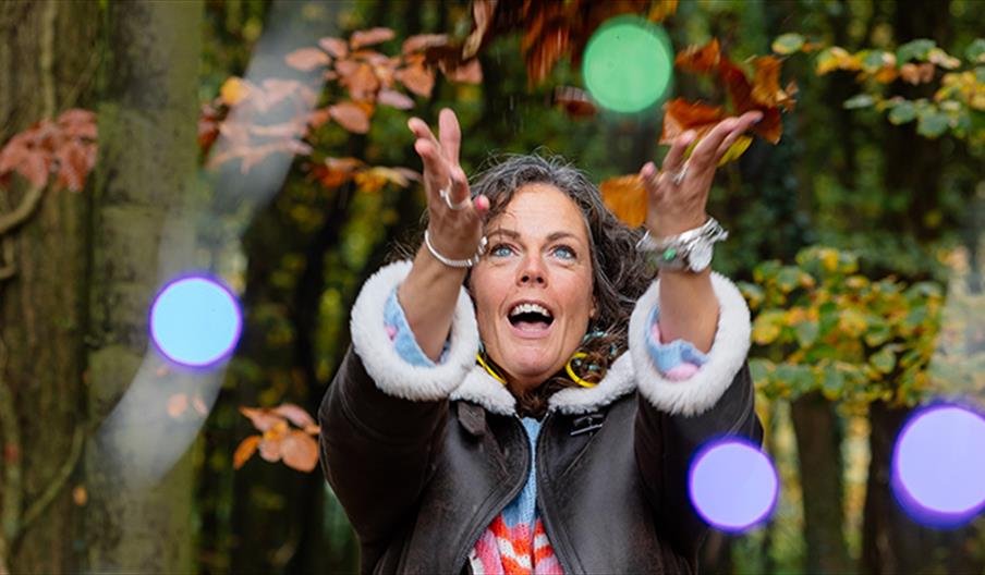 A woman standing in a forest and throwing multicoloured orbs in the air with a joyful expression on her face