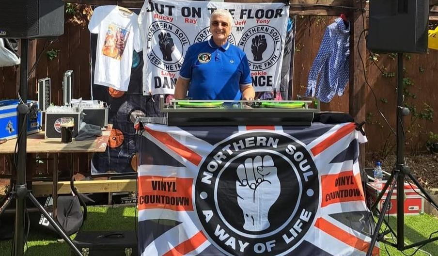 A man in a blue polo shirt standing behind a large record deck with a banner hung over it that reads; "Northern Soul: way of life"