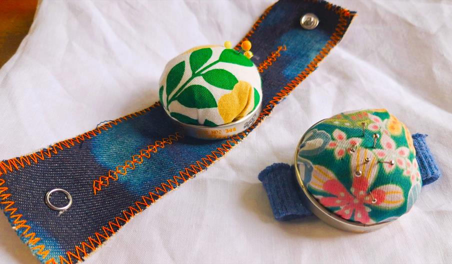 Two pin cushions made from multicoloured floral patterned fabric on blue demin wrist straps.