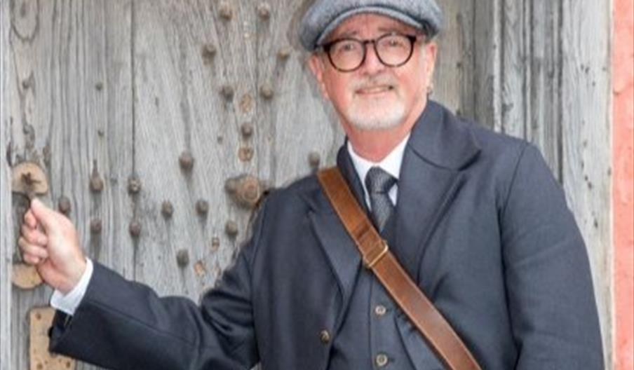 A smiling man in an Edwardian costume with flat cap and waistcoat opening a large old oak door