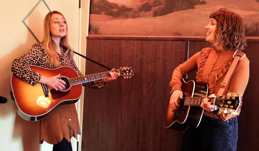 Two young women standing indoors playing acoustic guitars and singing while looking at each other