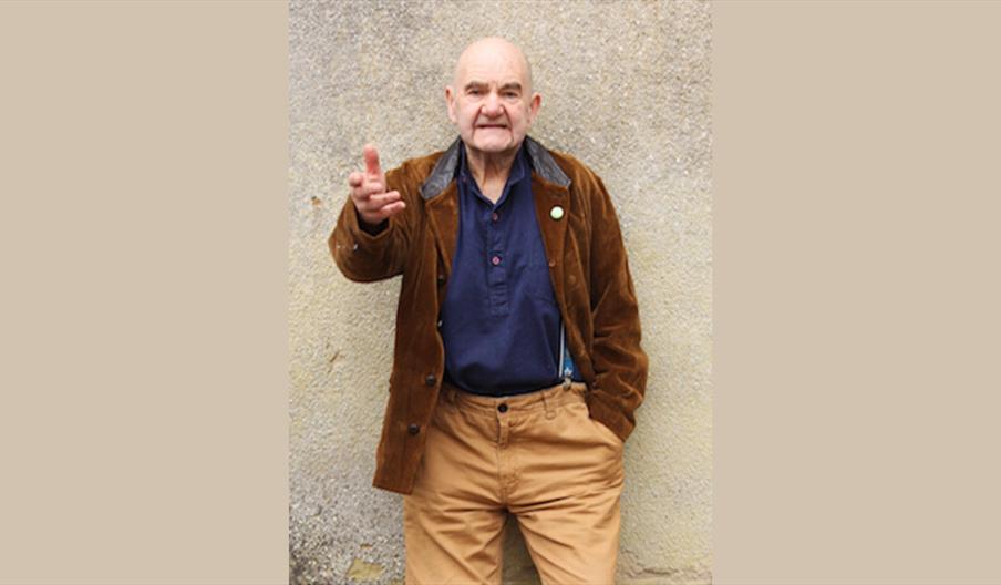 A man standing against a beige pebble dash wall with his hand out in mid speech